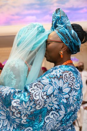 Traditional Nigerian wedding attire featuring intricate blue lace and headwrap against a purple sunset sky.