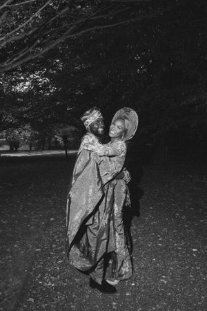 A black and white photo shows two people in traditional dress embracing under trees.