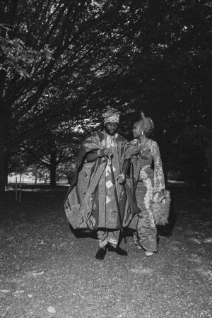 Black and white portrait of two people in traditional African formal wear standing outdoors.