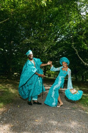 Two people in bright turquoise traditional Nigerian outfits dancing joyfully in a park.