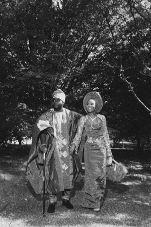 Black and white image of two people in traditional African formal wear standing together outdoors.