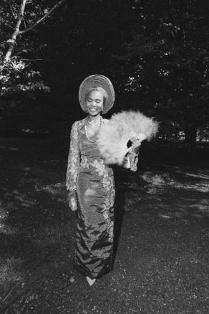 A woman in vintage dress and hat holds a feathered fan while posing outdoors in a black and white photograph.