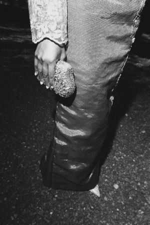 Close-up black and white photo of an embellished dress hem detail and hand holding a purse.