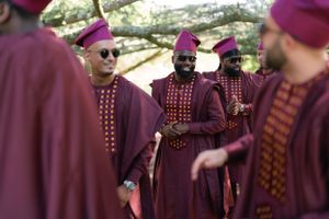 Group in burgundy ceremonial attire with magenta caps gathered under trees.