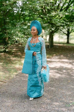 Woman in traditional Nigerian attire wearing turquoise gele and lace outfit with matching accessories.