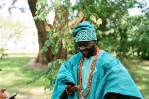 Person wearing traditional turquoise Nigerian agbada and cap while looking at mobile phone.