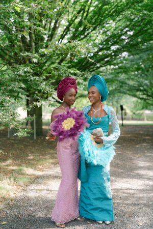 Two women in traditional Nigerian attire with gele headwraps in pink and turquoise colors.