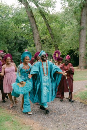 Wedding party wearing vibrant turquoise and burgundy traditional African attire walking joyfully down a garden path.