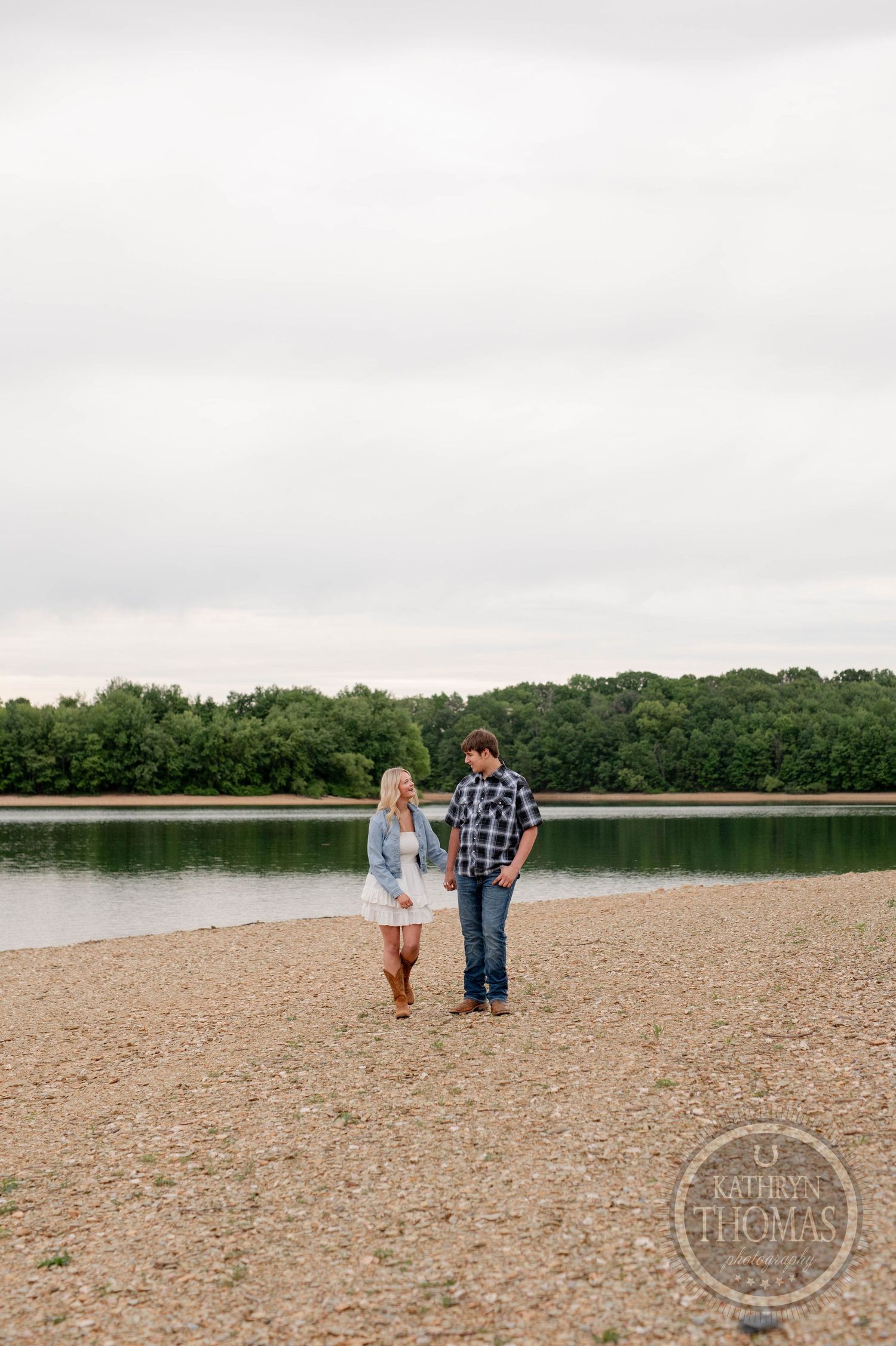 Couple walking together on a wide rocky beach surrounded by green trees along the shoreline.