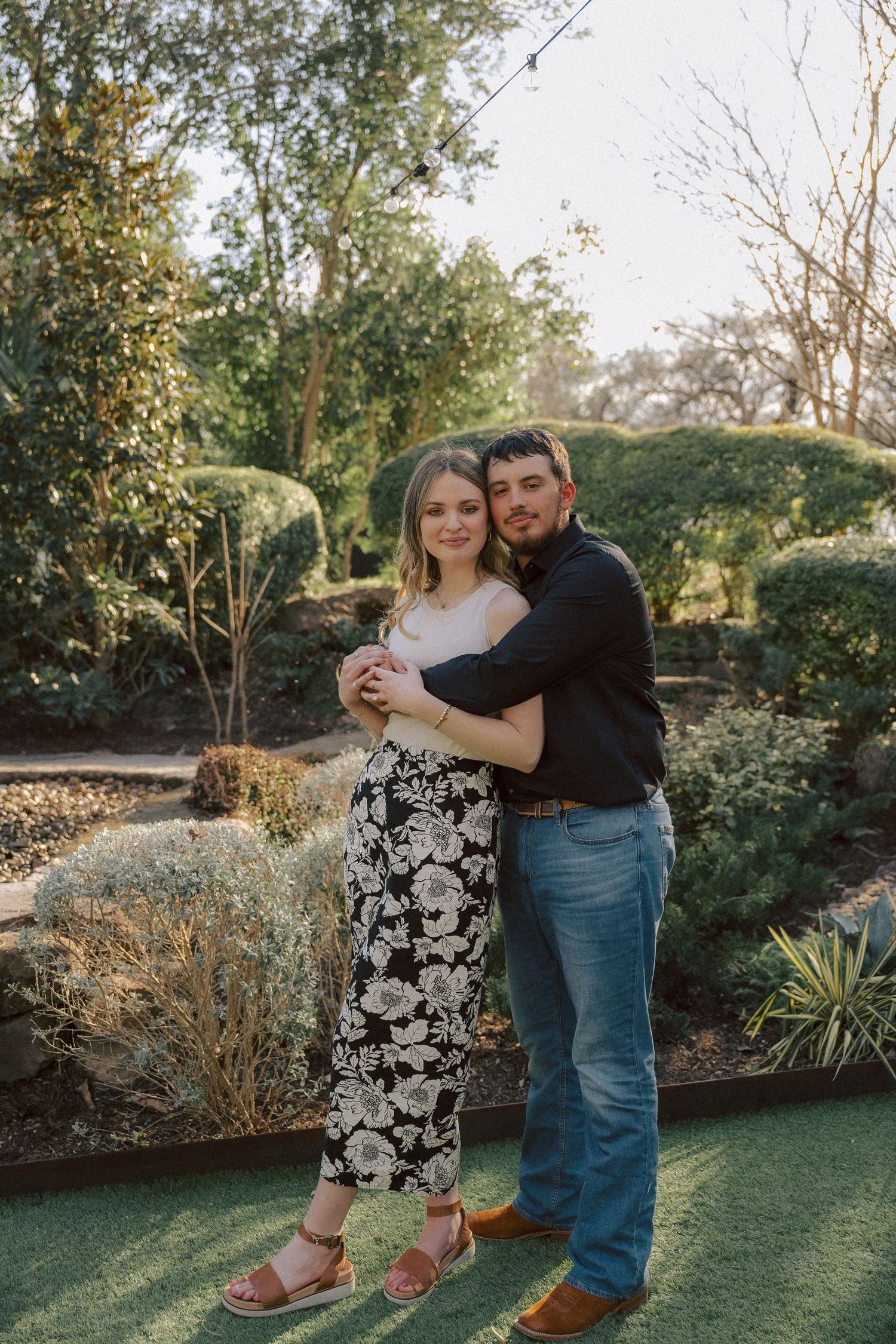 A couple embraces while standing in a garden setting with green landscaping and trees during golden hour lighting.