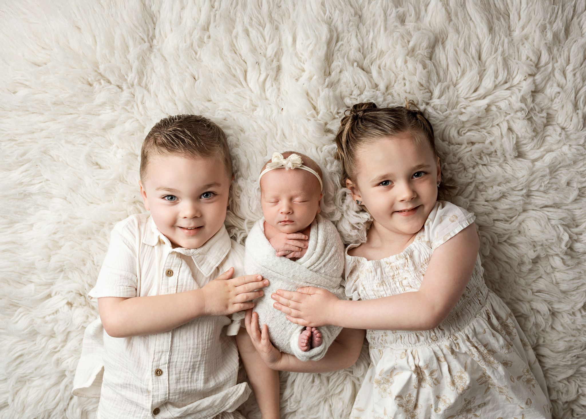 Two children in white outfits lovingly embrace a newborn baby on a soft cream-colored blanket.