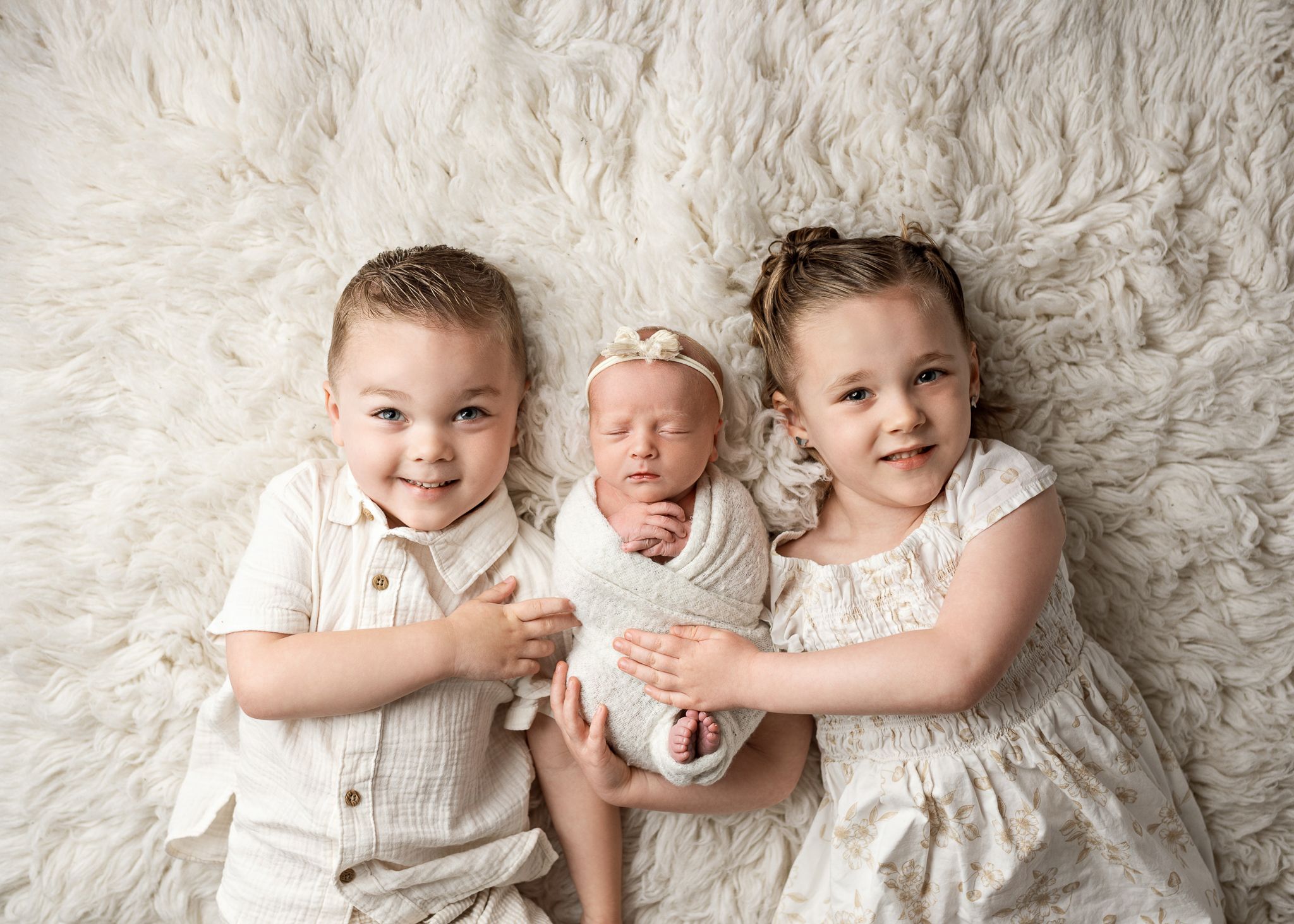 Three young siblings in white outfits pose together on a plush cream-colored fur blanket for a heartwarming portrait.