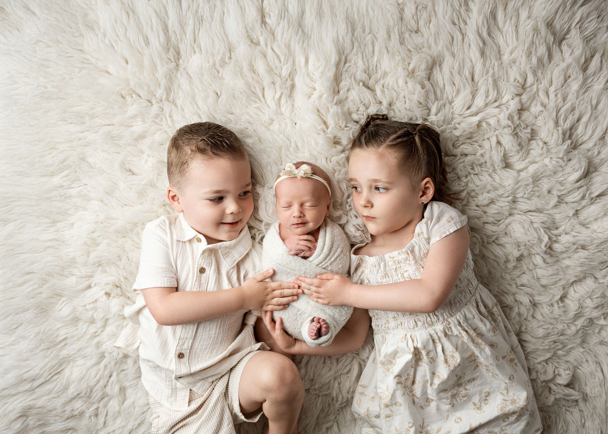 Three children in white outfits lying together on a plush white fur rug during a newborn photo session.