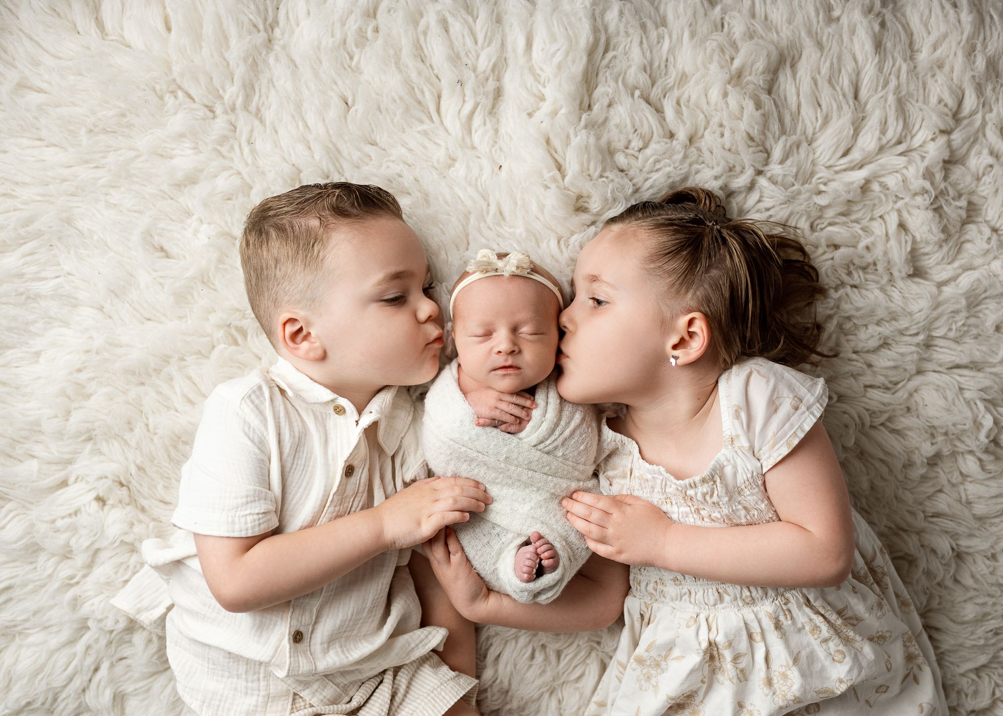 Three siblings share a tender moment on white fur bedding in matching light-colored outfits.