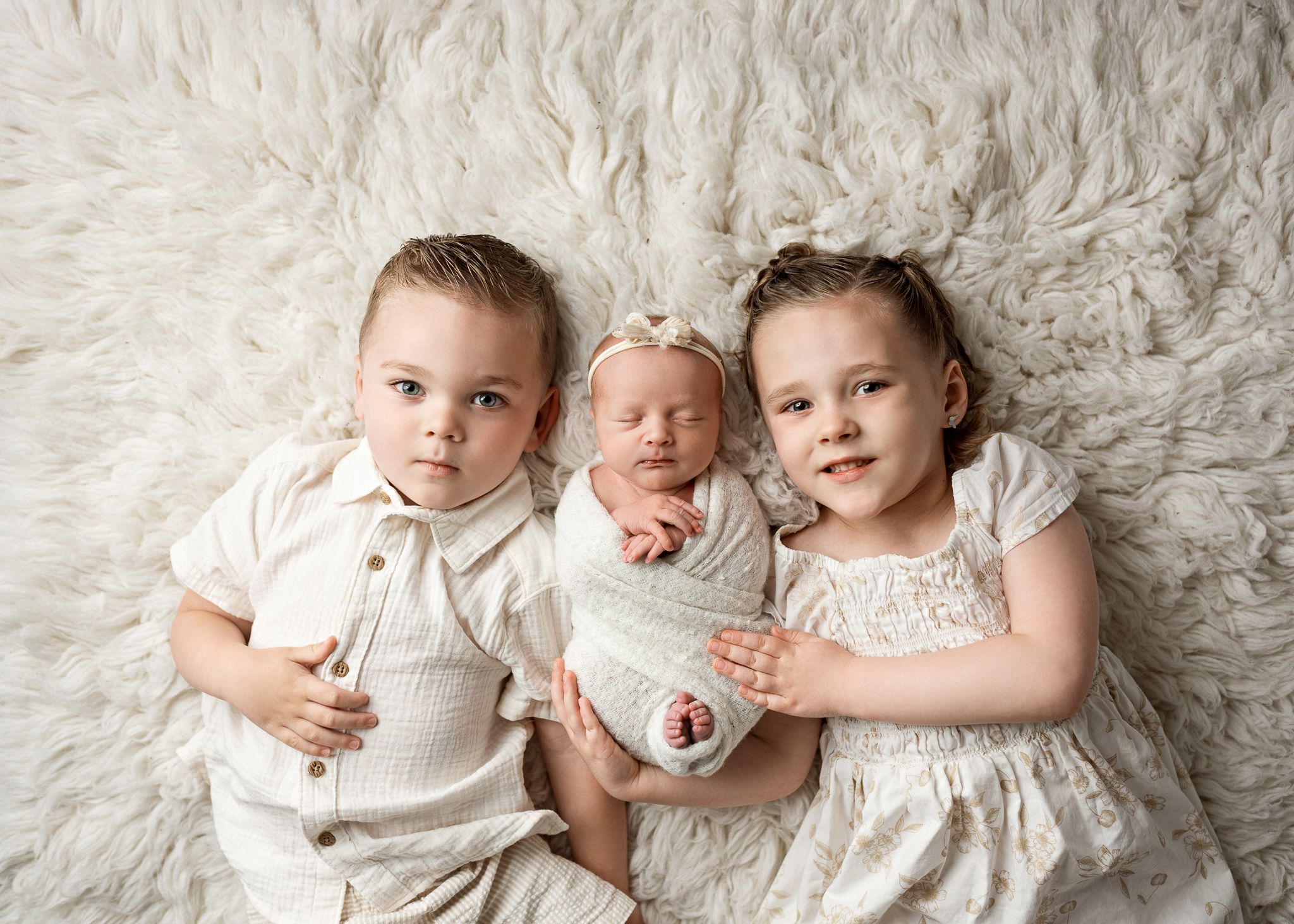 Three siblings posed together on a plush white fur backdrop wearing coordinated cream-colored outfits.