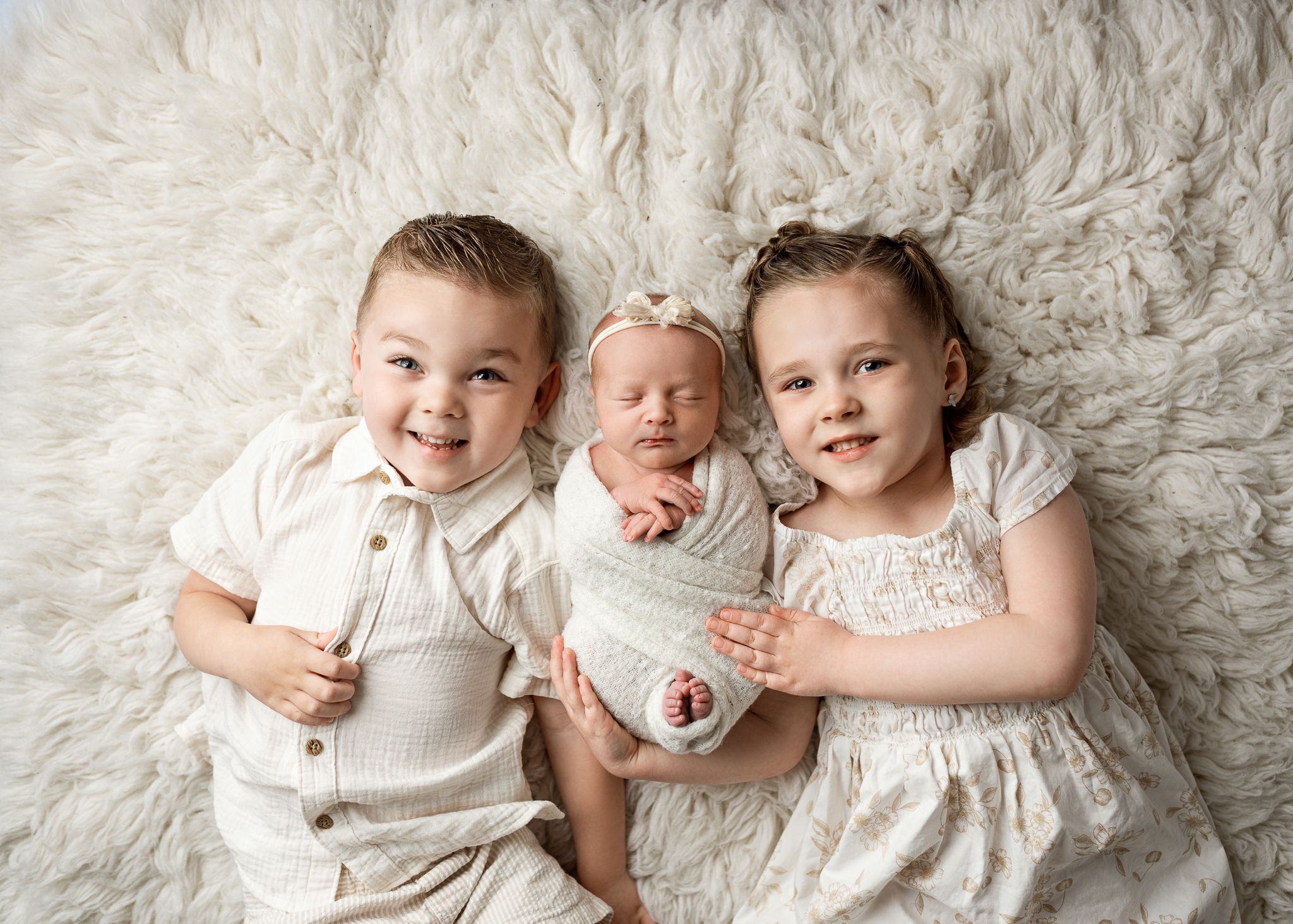 Three children in cream outfits smiling while posed together on a fluffy white background.