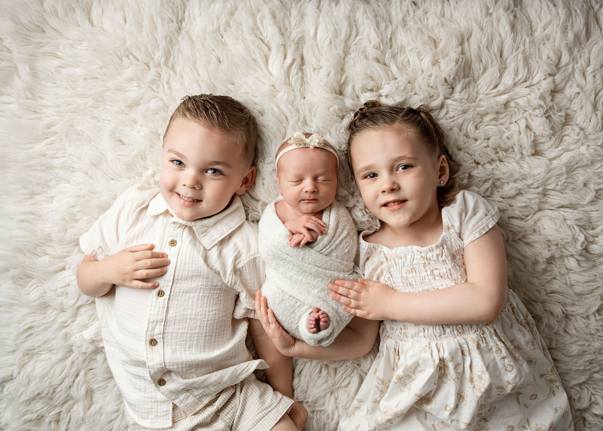Three siblings in light colored outfits pose together on a soft white fur rug for a professional portrait.