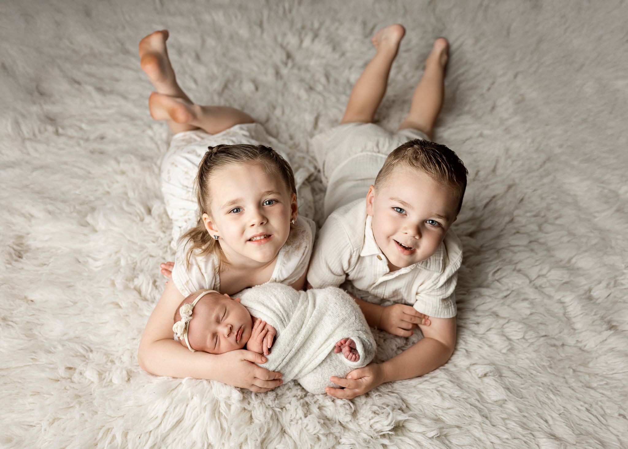 Three young children laying together on a white fuzzy rug, wearing light colored outfits.