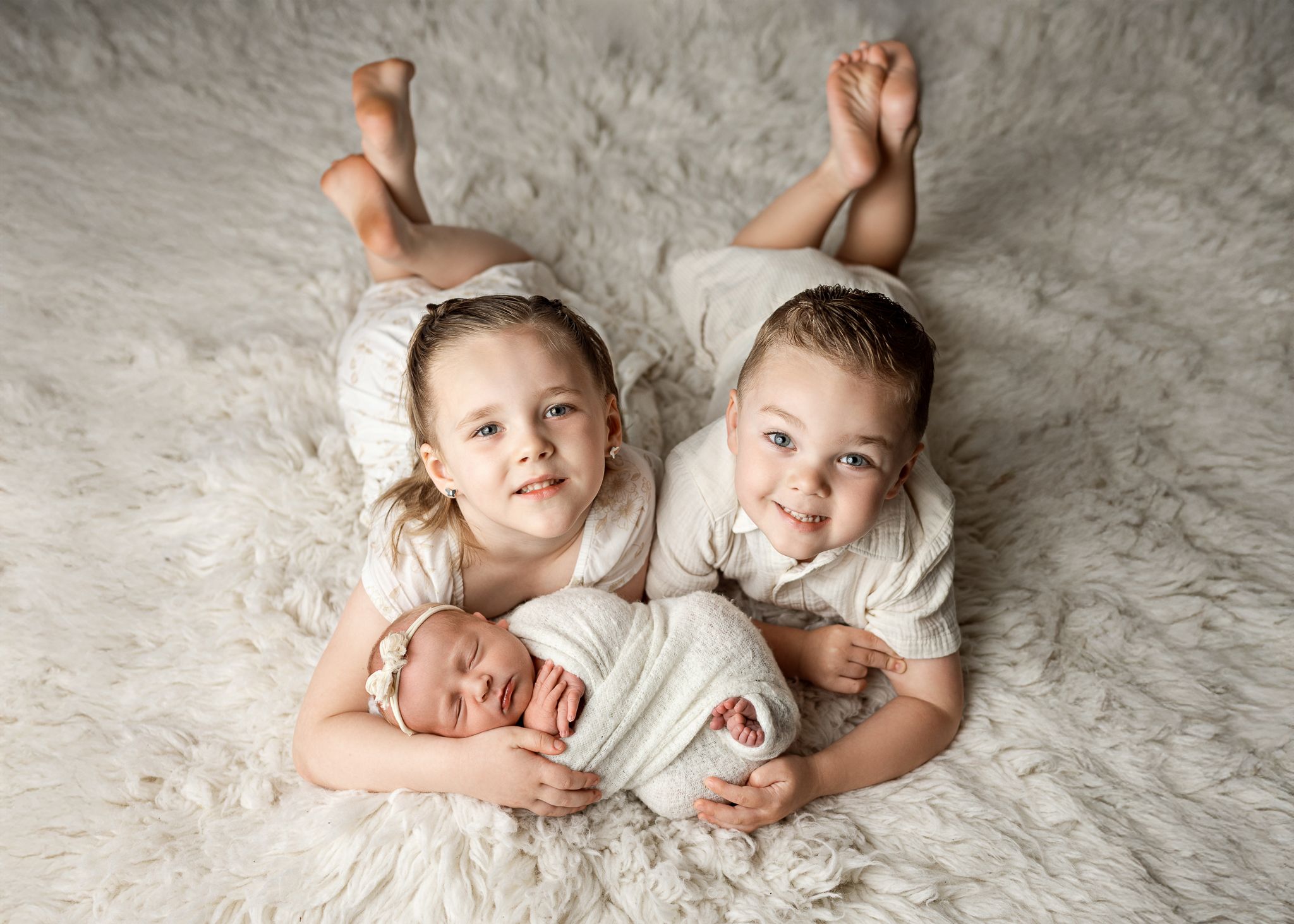 Three young children wearing white outfits lying together on a soft gray textured rug while looking up at camera.