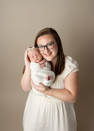 A tender portrait showing a newborn baby wrapped in white swaddling being held close against a neutral beige background.