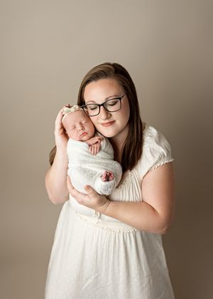 A tender moment between a woman in glasses and white dress holding a sleeping newborn baby swaddled in white.