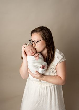 A woman in a white dress tenderly embraces her sleeping newborn baby wrapped in a soft blanket against a neutral backdrop.