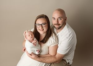 A couple in white clothing poses together while holding a baby in a studio setting.
