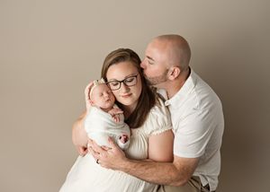 A family portrait in neutral colors shows parents embracing their baby against a beige studio backdrop.