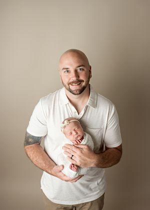 A gentle portrait of an adult in a white polo shirt tenderly holding a sleeping newborn baby against a neutral background.