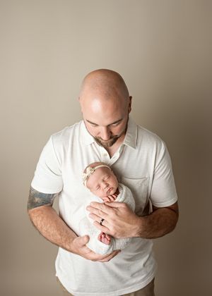 An intimate moment captured as an adult in white gazes down lovingly at a sleeping newborn baby against a beige backdrop.