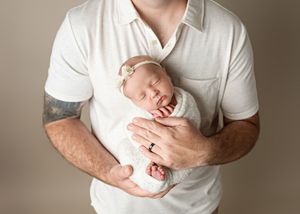 A sleeping newborn baby wearing white is cradled securely against a white-shirted chest with tattooed arms.
