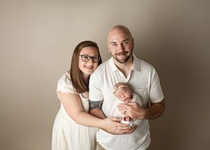 Family portrait with a couple in white clothing holding their newborn baby against a neutral backdrop.