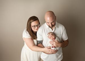 A couple in white outfits tenderly holds and gazes at their newborn baby against a neutral beige backdrop.