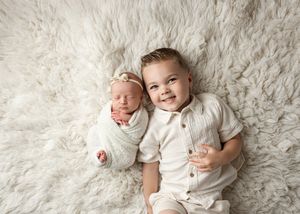 Two children in white outfits laying on a soft white fur background for a studio portrait session.