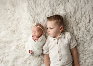 A young child in white clothing laying next to a swaddled newborn on a plush white fur backdrop.