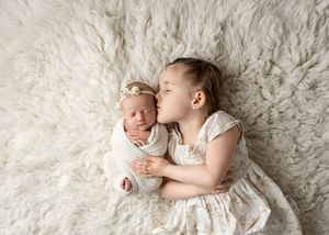 Two children in white lace dresses lying on a fluffy cream-colored rug while sharing a tender moment together.