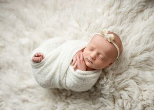 A sleeping newborn wrapped in a soft white swaddle lies peacefully on a fluffy cream-colored blanket.