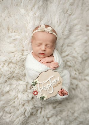 Sleeping newborn wrapped in white blanket lying on fluffy white fur background with delicate floral decorations.