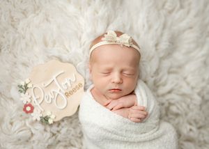 A sleeping newborn wrapped in white swaddling lies on soft fur bedding with a floral decorative sign beside them.