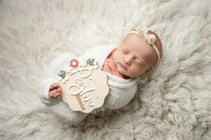 Newborn baby sleeping peacefully on soft white fur blanket wearing knit cap and headband with floral accents.