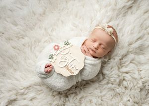 A sleeping newborn swaddled in white fabric rests on a fluffy cream-colored fur blanket with a wooden milestone marker nearby.