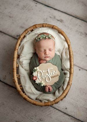 A peaceful sleeping newborn in sage green wrap lies in a woven bamboo basket with cream bedding and floral accents.