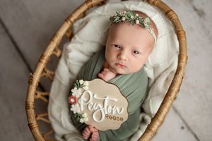 A newborn baby swaddled in soft sage green fabric rests peacefully in a woven rattan basket during a professional photoshoot.