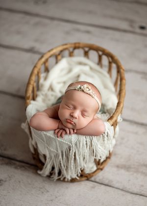 A sleeping newborn nestled in a white blanket inside a round woven basket on wooden floorboards.