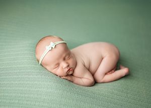 Sleeping newborn baby curled up peacefully on soft green fabric wearing delicate white headband.