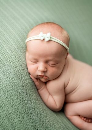 Peaceful newborn resting on sage green textured surface wearing delicate white bow headband.