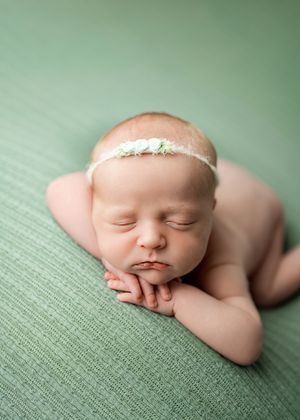 Sleeping newborn wearing delicate floral headband rests peacefully on sage green textured fabric.