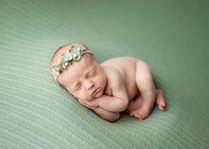 Peaceful sleeping newborn wearing delicate floral headband curled up on soft green textured blanket.