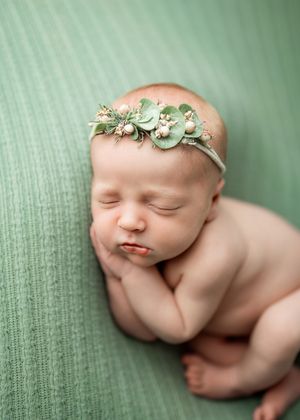 Sleeping newborn wearing mint green floral headband rests peacefully on sage green textured blanket.