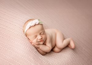 A peaceful sleeping newborn rests on a pink textured blanket wearing a delicate lace headband.